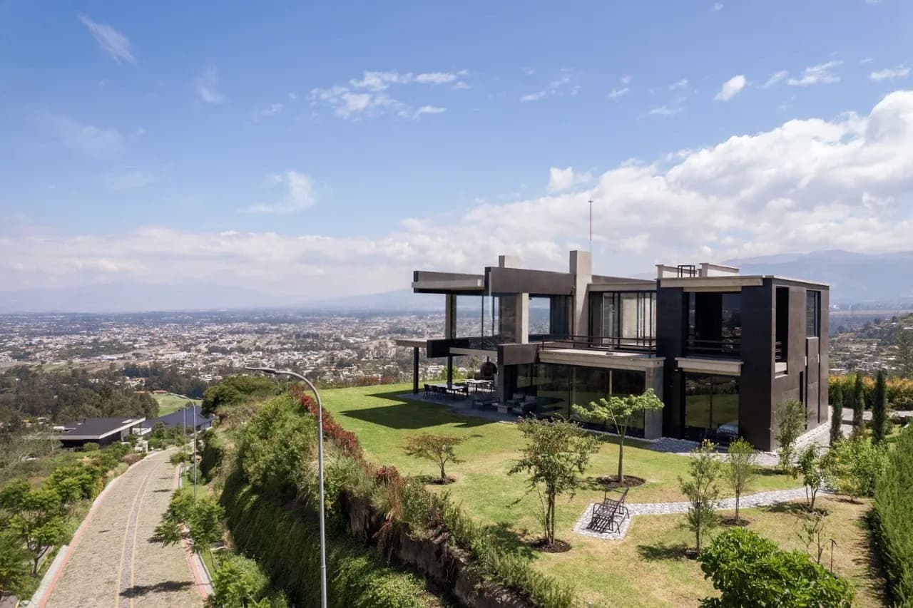 90 Degree House sobre una ladera del Ilaló, con volúmenes oscuros y grandes vidrios, un jardín que llega al borde del talud y el valle de Quito al fondo